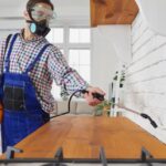 A person wearing a protective mask, goggles, and overalls demonstrates the termite treatment process by spraying pesticide on a wooden kitchen countertop near a white brick wall.