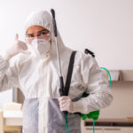 A person wearing a full-body protective suit, gloves, and a mask stands indoors holding spraying equipment and making a phone call gesture—ready to deliver year-round pest control services for complete home protection.