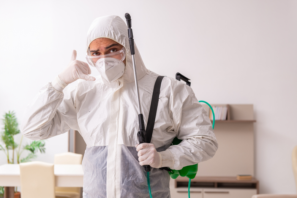 A person wearing a full-body protective suit, gloves, and a mask stands indoors holding spraying equipment and making a phone call gesture—ready to deliver year-round pest control services for complete home protection.