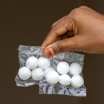A hand is holding two small clear plastic bags, each containing five round white mothballs, perfect for home protection, against a plain dark background.
