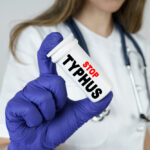 A person wearing a white medical coat and blue gloves holds a container labeled “STOP TYPHUS,” promoting typhus prevention. A stethoscope hangs around their neck, highlighting their role as a healthcare professional.