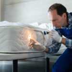 A man wearing gloves and overalls inspects a mattress with a flashlight, focusing on dark spots along the side, suggesting a bed bug exterminator performing pest control in a bedroom setting.