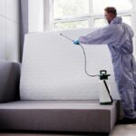 A person in protective gear and gloves uses a spray canister to disinfect a mattress propped against a gray couch near a window, following expert Bed Bug Treatments for a cleaner, bug-free home.