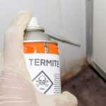 A gloved hand holds a can of Pest Control termite spray labeled "TOXIC" with a skull and crossbones symbol, aimed at the corner where the tiled wall meets the floor.