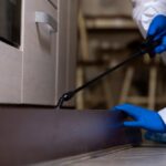 A person wearing blue gloves and protective clothing uses a spray nozzle to apply safe flea solutions along the baseboard of a kitchen cabinet for effective pest control.