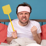 A man sits in bed wearing a pink sleep mask, clenching his fist and holding a yellow fly swatter with a frustrated expression—clearly in need of a Bed Bug Elimination solution. Coral pillows and a white blanket stand out against the blue background.