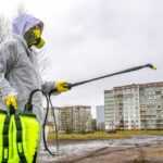 A person wearing protective gear and a gas mask sprays disinfectant from a large yellow container, providing home flea control in an outdoor area near apartment buildings on a cloudy day.