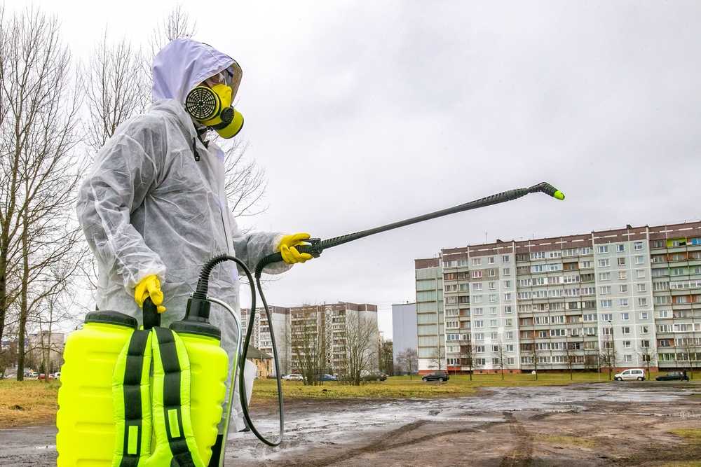 A person wearing protective gear and a gas mask sprays disinfectant from a large yellow container, providing home flea control in an outdoor area near apartment buildings on a cloudy day.