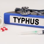 A binder labeled "Typhus Disease" rests on a white surface with a stethoscope, syringe, and a tipped-over pill bottle spilling red-and-white capsules, highlighting the focus on symptoms and treatment.