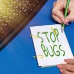 A person writes "STOP BUGS" on a notepad with a green pencil, next to a black keyboard on a blue desk. Yellow paperclips secure the notepad. Sunlight shines in as they consult a Bed Bug Guide for tips on achieving a Pest-Free Home.