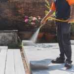 A person wearing a short-sleeve orange shirt and dark pants uses a garden hose to spray water on a concrete patio, keeping the area tidy as part of a Residential Pest Control routine. Potted plants and a brick wall are in the background.