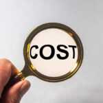 A hand holding a magnifying glass enlarges the word "COST," symbolizing a close examination of termite exterminator cost on a white background.