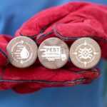 A person wearing bright red gloves holds three wooden discs, each with a white icon and text representing water treatment, Safe Pest Control, and sanitation. The background is blurred.