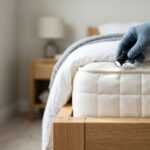A person wearing a blue glove shines a flashlight on the edge of a mattress, possibly inspecting for pests in a neatly made bedroom with light-colored furniture—an important step before considering bed bug extermination cost.